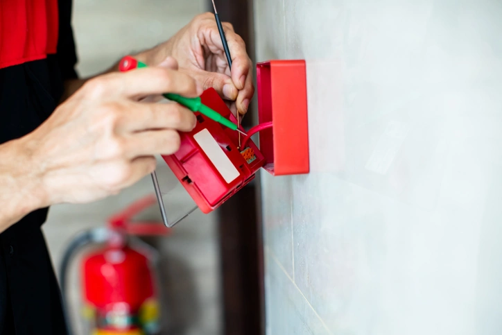 Close-up of technician hands installing and wiring a red fire alarm manual call point box on a wall